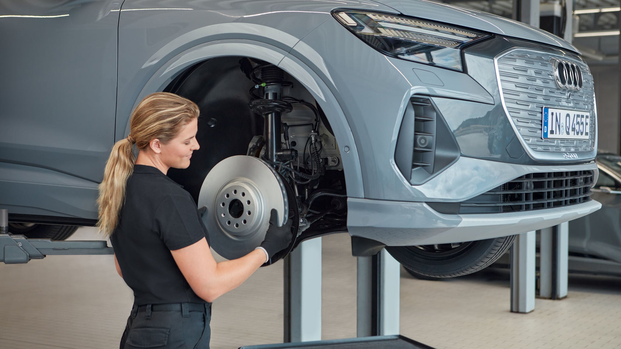 Eine Mechanikerin prüft die Bremsanlage eines angehobenen grauen Audi-Fahrzeugs in einer Werkstattumgebung.