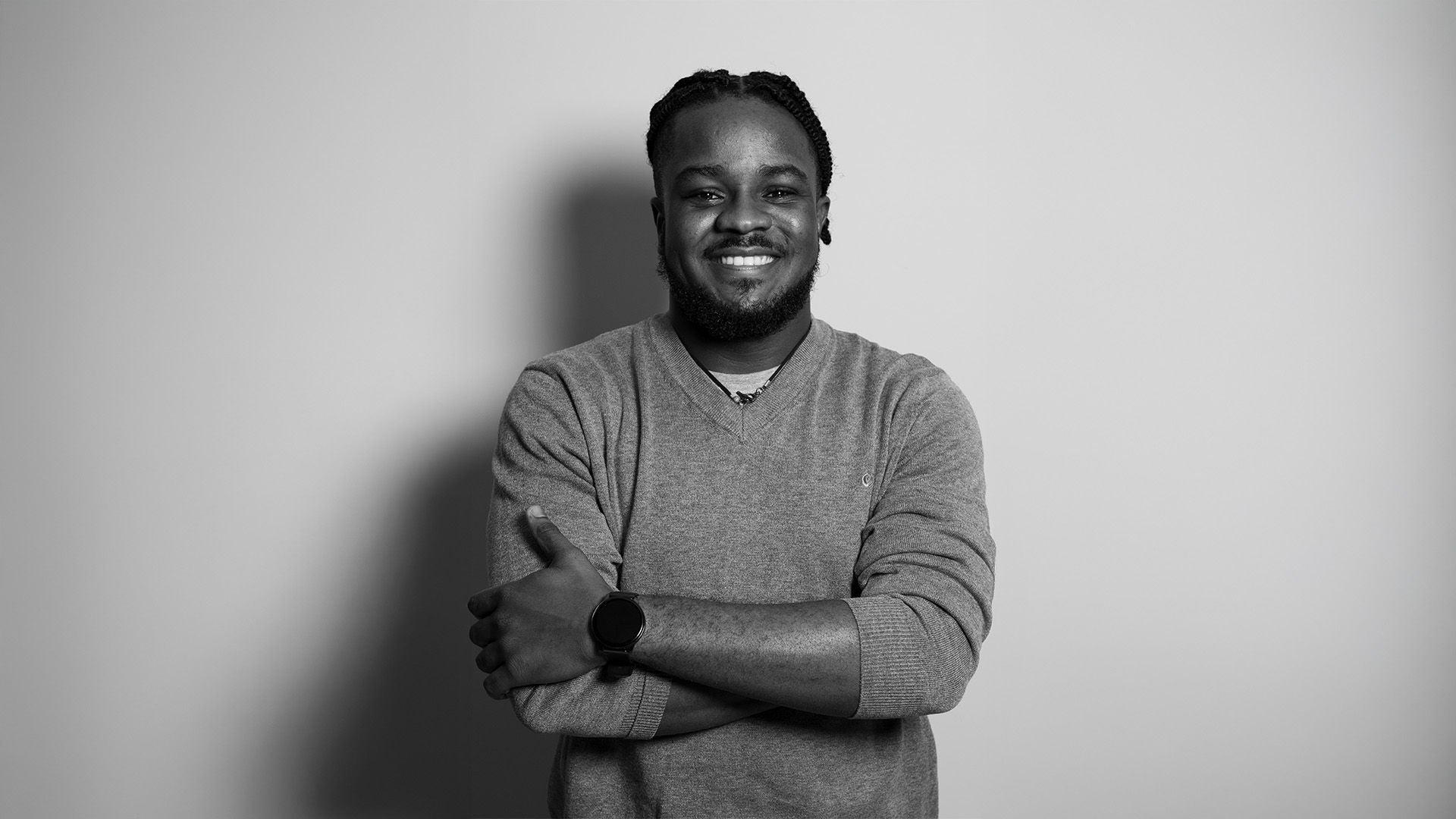 Schwarz-weiß Porträt eines Mannes im grauen Pullover mit Uhr, lächelnd, vor hellem Hintergrund.