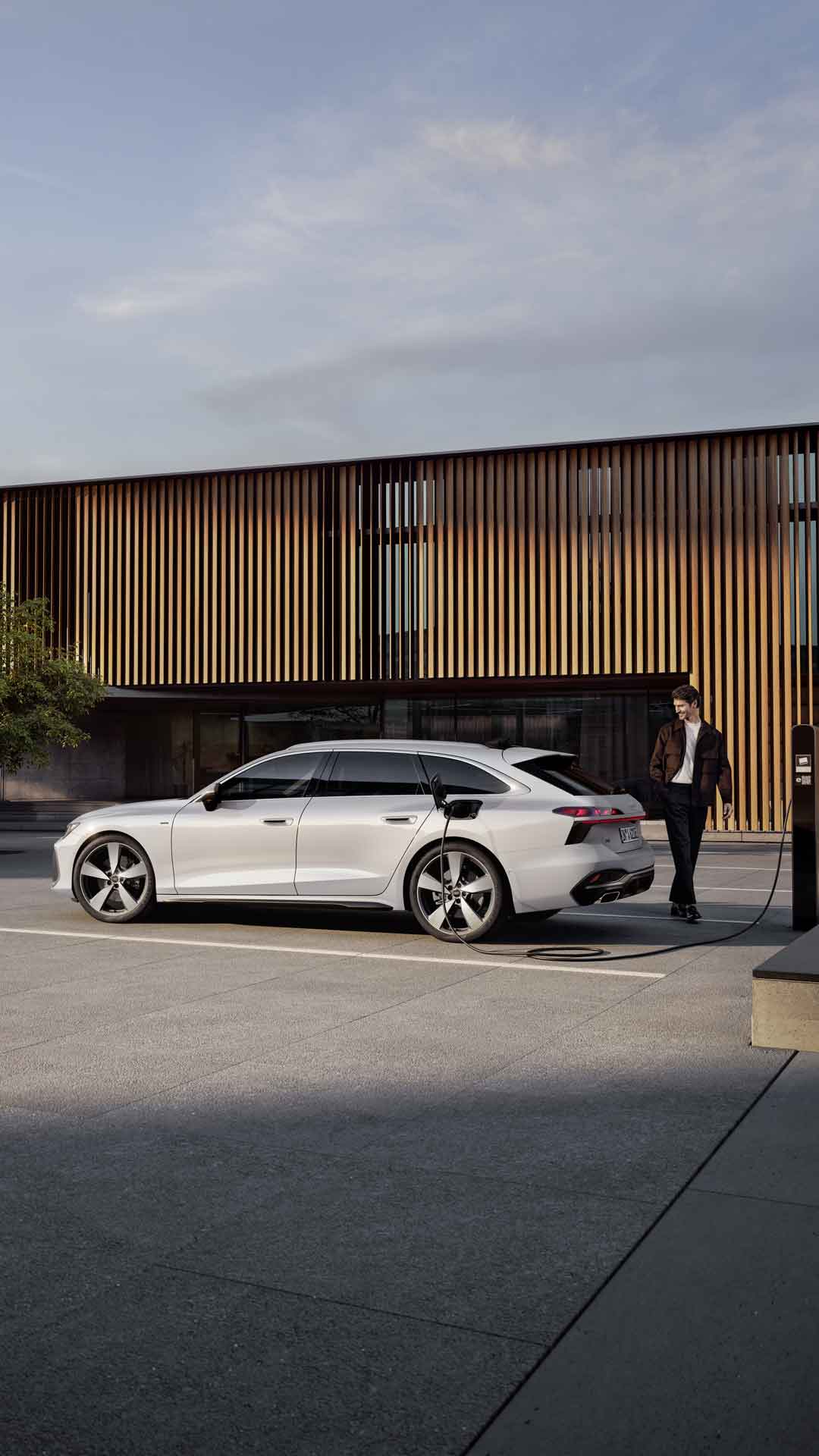 A6 Avant e-hybrid står parkerad vid sidan av entrén till en stor byggnad och laddas via en strömkabel. En medelålders man står bakom bilen.