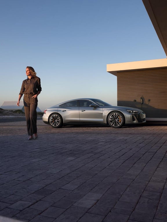 Eine Frau in eleganter, dunkler Kleidung geht an einem silbernen Audi S e-tron GT vorbei, der an einer modernen Ladestation vor einem stilvollen Gebäude angeschlossen ist.
