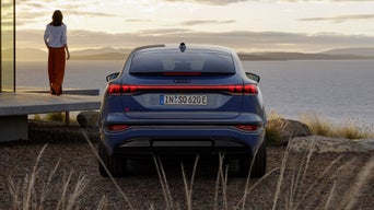 Der SQ6 Sportback im Heckprofil. Das Fahrzeug steht neben der Veranda eines Hauses, auf der eine Frau steht. Die Frau und das Fahrzeug sind einem großen Gewässer zugewandt.