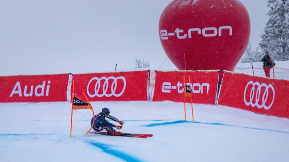 Ein Skifahrer in der Kurve. Im Hintergrund rote Banden mit dem Audi-Logo und dem Schriftzug e-tron sowie ein roter Ballon mit dem Schriftzug e-tron.