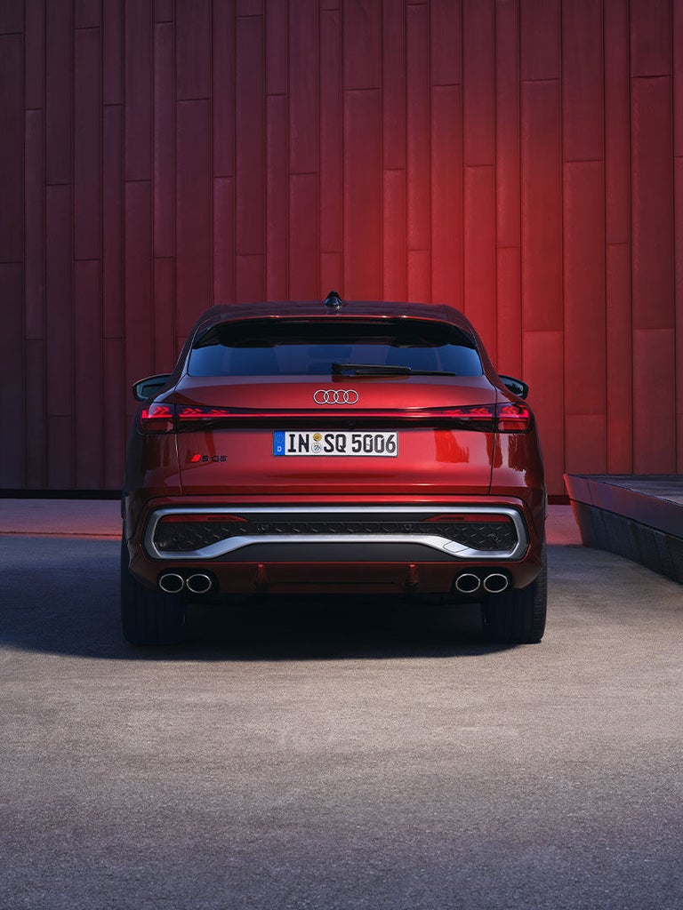 Heckansicht eines roten Audi SQ5 Sportback mit vier Auspuffrohren und Modellschriftzug vor moderner Fassade in dunkler Umgebung.