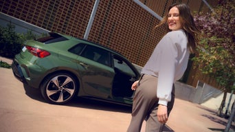 Lächelnde Frau blickt zurück, während sie neben einem grünen Audi A3 mit geöffneter Tür auf einem sonnigen Gehweg steht.