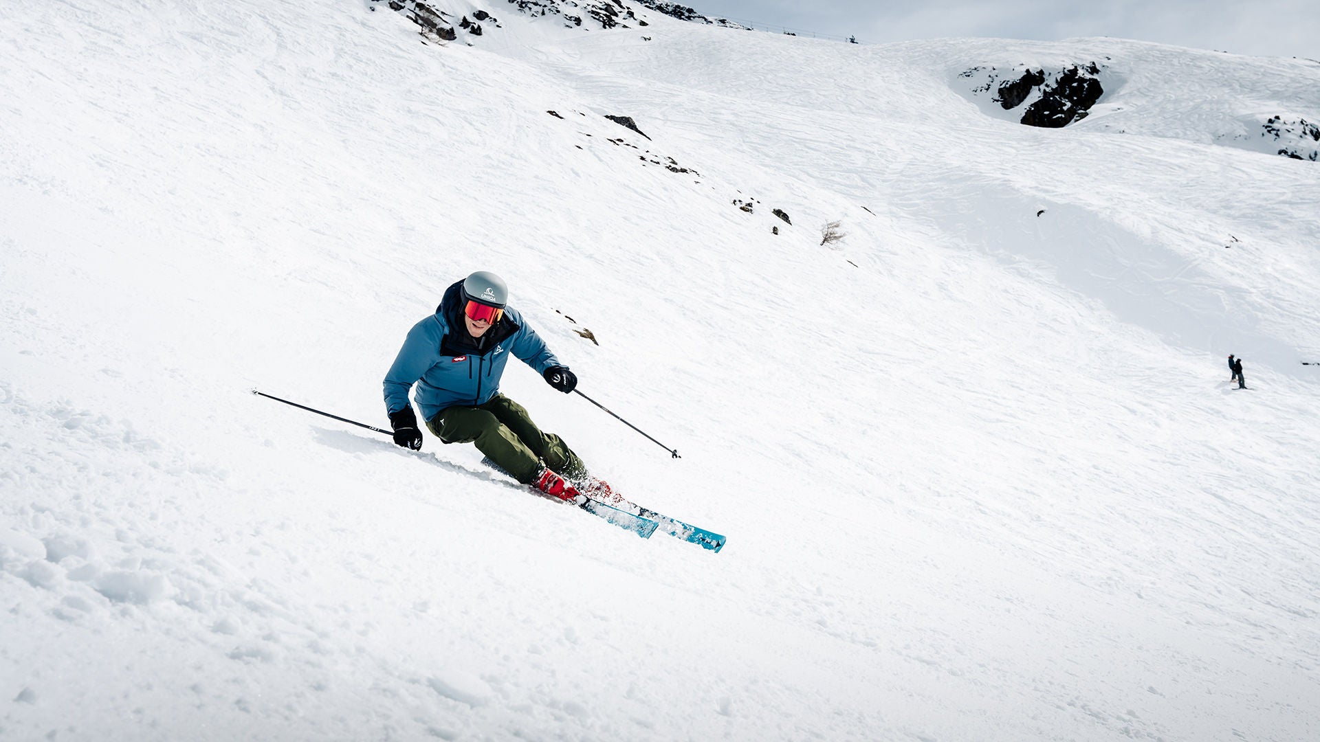 Skier in blue jacket turns in deep snow on a slope; red ski boots and ski poles visible.