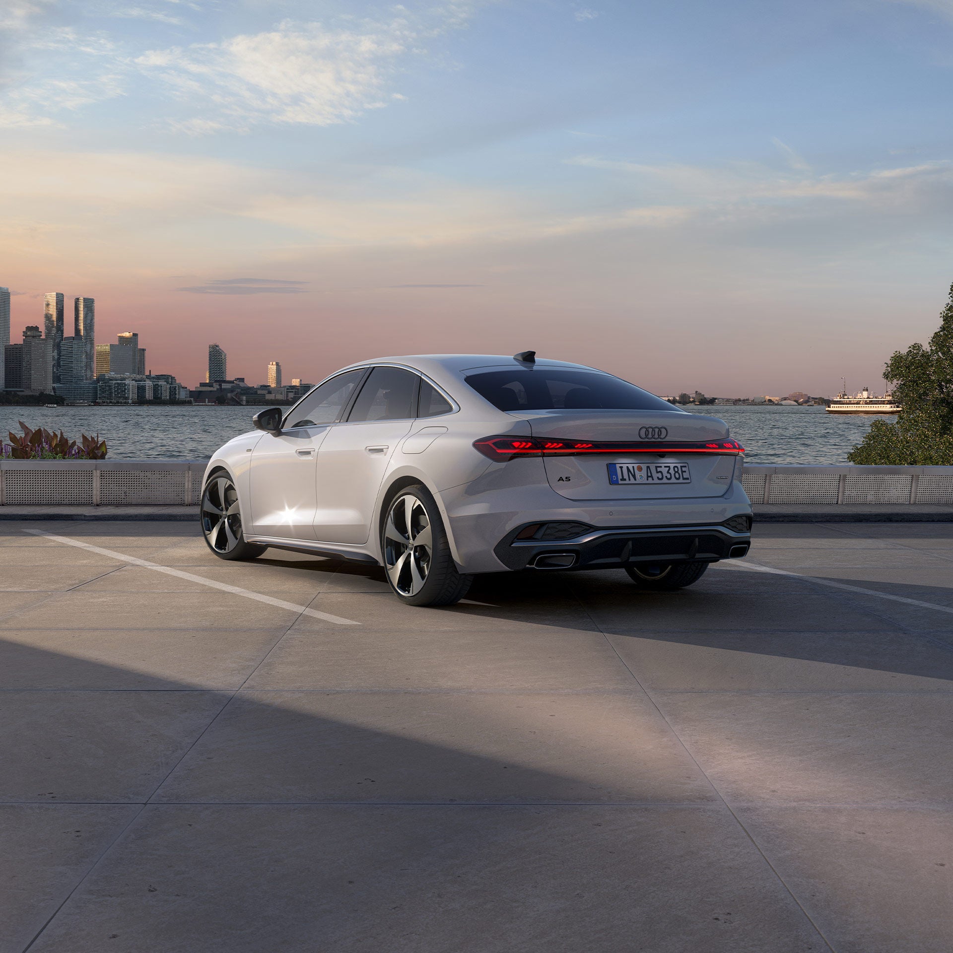 Die A5 Limousine e-hybrid steht im schrägen Heckprofil auf einem Parkplatz am Ufer eines Sees. Dahinter ist die Skyline einer Stadt bei Dämmerung zu sehen.
