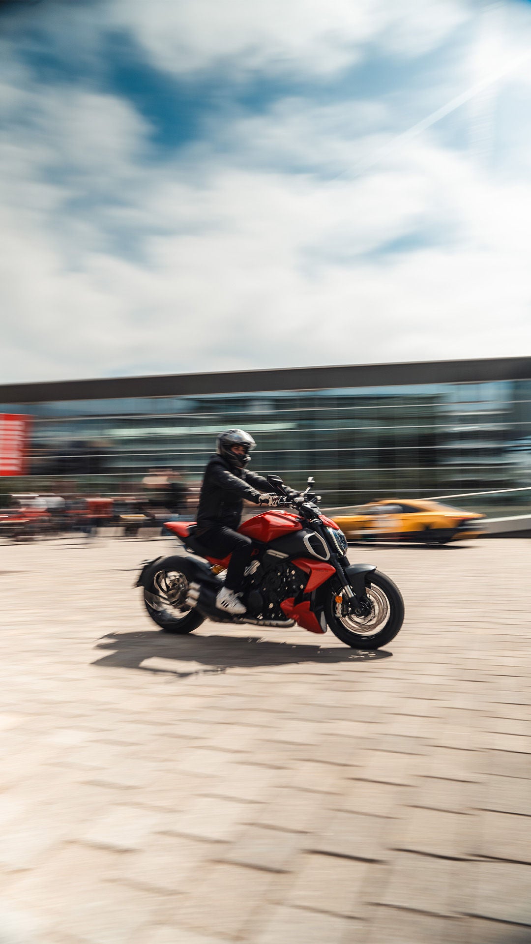 Person mit Helm fährt rotes Motorrad über Platz, modernes Gebäude im Hintergrund.