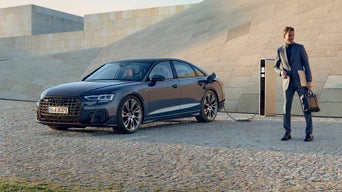 Ein dunkelblauer Audi A8 TFSI e lädt an einer Ladesäule, ein Mann in grauem Anzug steht mit Aktentasche und Laptop in der Hand daneben.