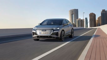 An Audi Q4 Sportback e-tron cruising on a modern highway with a city skyline in the background under a clear sky.