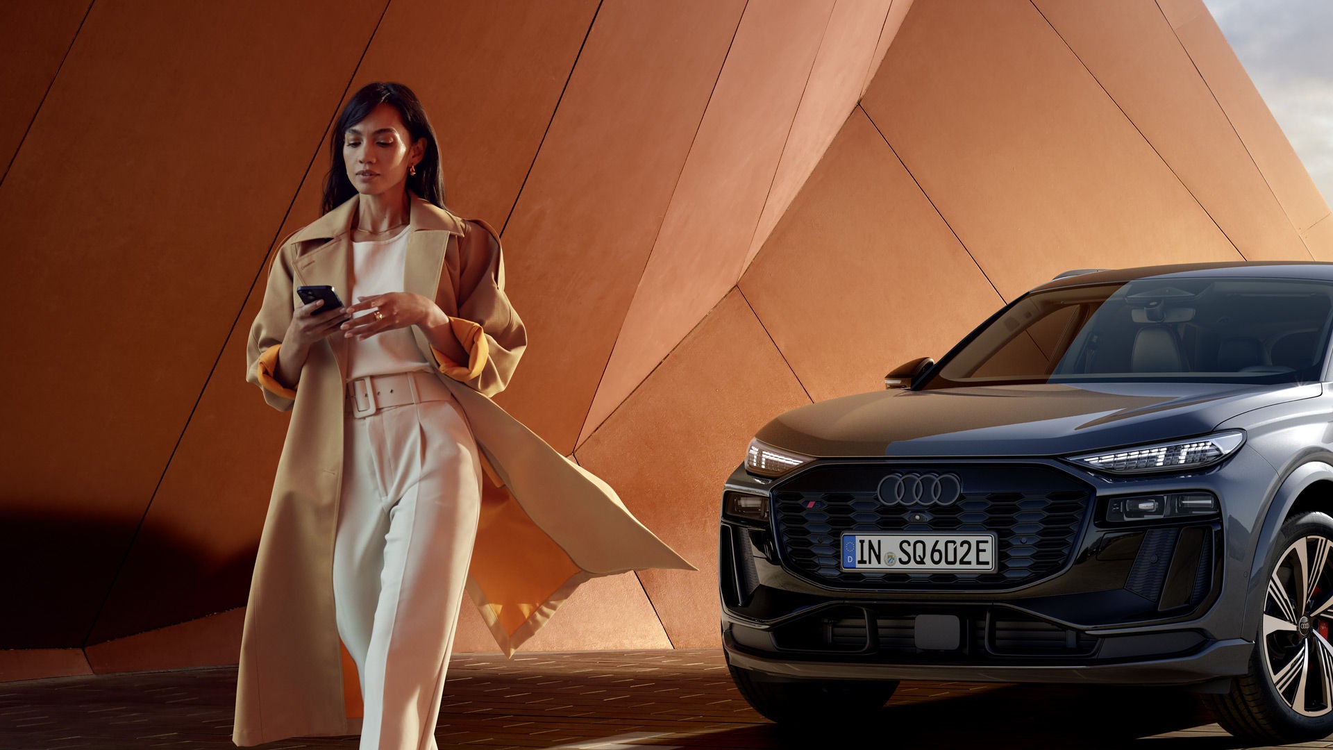 Frau steht lächelnd vor zwei geparkten Audi-Fahrzeugen und bedient ein Smartphone, moderner Holzbau im Hintergrund.