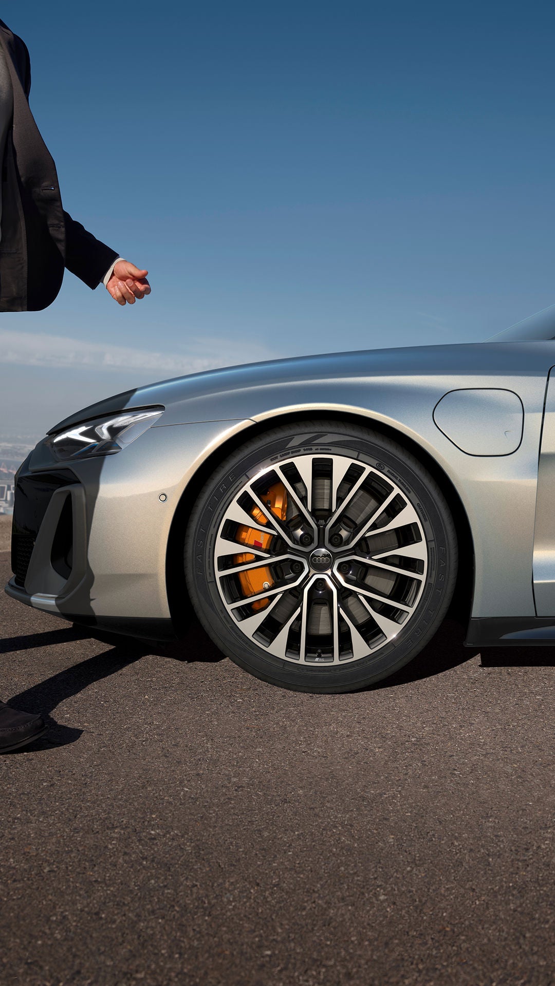 A man in a suit walks beside a silver car, showcasing its stylish wheel and brake calipers against a clear sky backdrop.