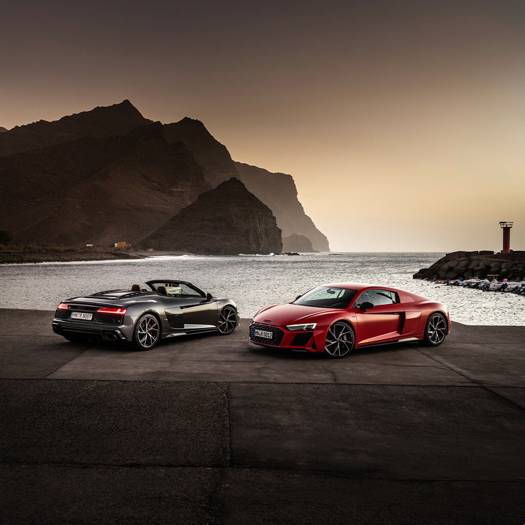 Zwei Sportwagen am Meer vor Felsküste bei Sonnenuntergang, ruhige Bucht und dramatische Bergkulisse im Hintergrund.