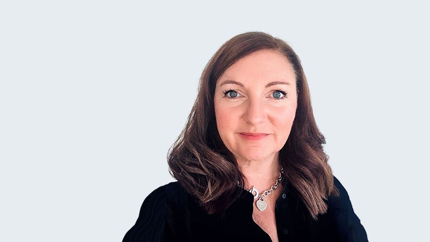 Antje Stephanie Ham: Porträt Frau mit brünettem, leicht gewellten Haar, silberner Kette und schwarzer Bluse vor grauem Hintergrund.