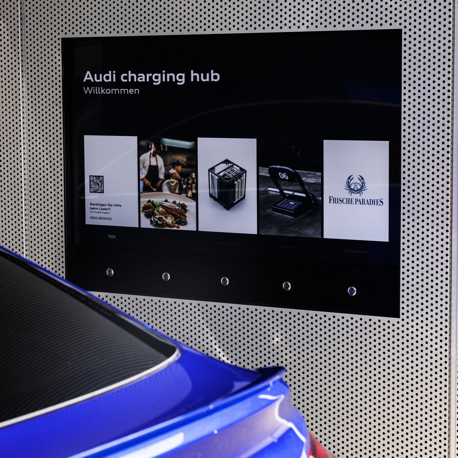Digitales Display mit der Aufschrift „Audi charging hub – Willkommen“, daneben fünf Info-Kacheln, vorne ein blaues Auto.