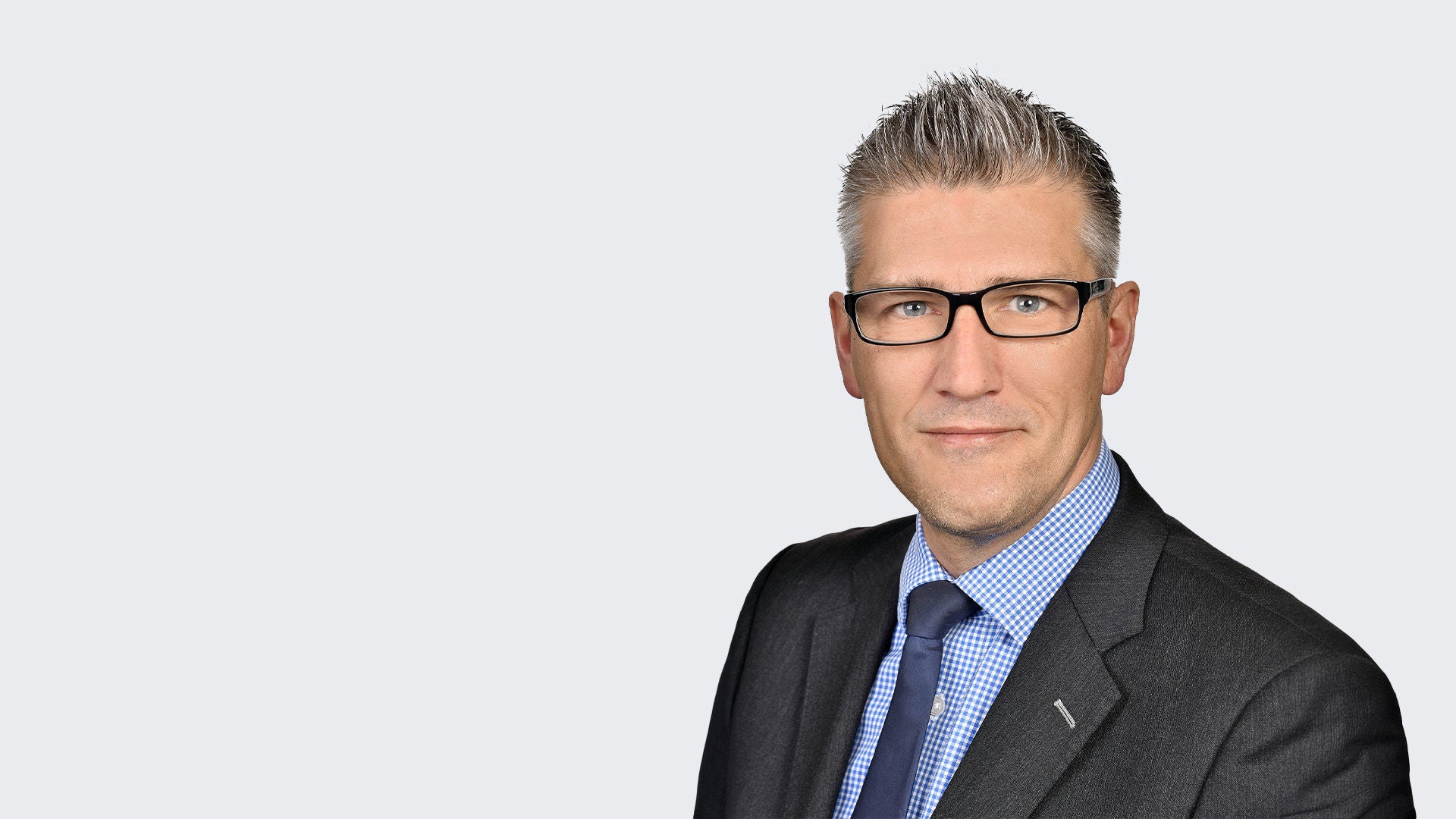 Mark Tworek: Porträt eines Mannes mit Brille im dunkelgrauen Anzug und blaukariertem Hemd vor hellgrauem Hintergrund.