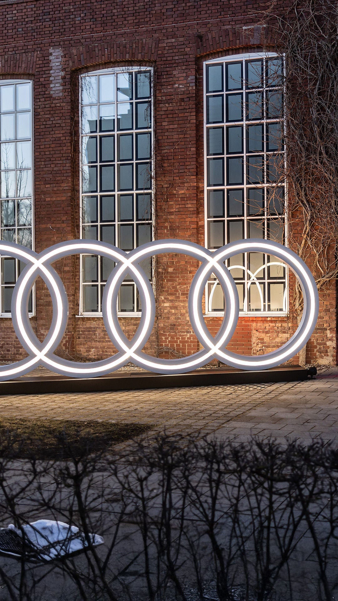 Im Hintergrund befindet sich ein altes Fabrik-Backsteingebäude mit hohen weissen Sprossenfenstern. Aus der geöffneten Tür dringt ein pinkfabener Lichtstrahl hervor. Im Vordergrungd befindet sich lebensgroße weiss leuchtende Audi Ringe.