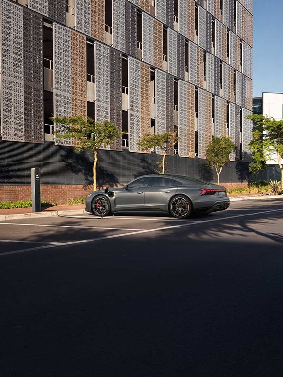 Ein Audi RS e-tron GT lädt an einer Straßenladestation vor einer modernen Fassade mit Bäumen und Schatten.