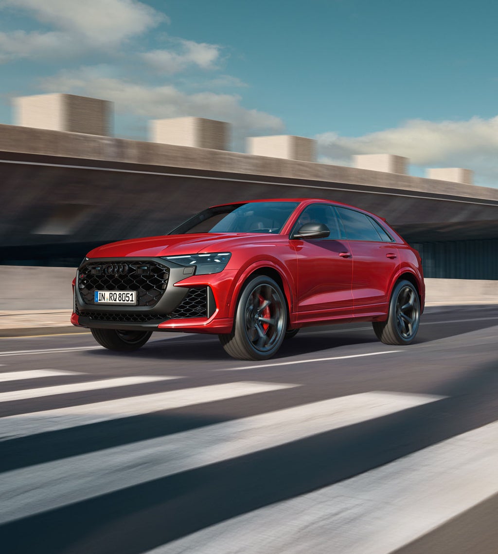 The RS Q8 SUV performance is approaching a crosswalk from the front at an angle. To its right stands a futuristic building with a concrete-look exterior.