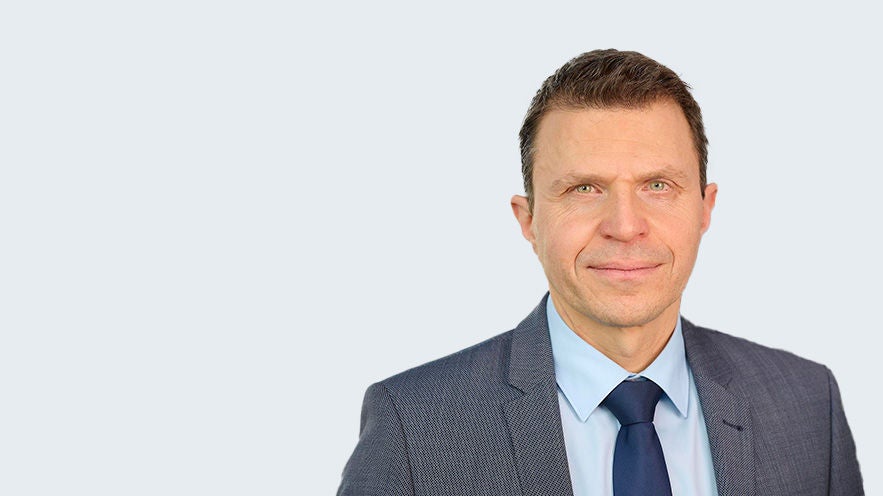 Stefan Trauf: Porträt eines älteren Mann im grau/blauen Anzug vor grauem Hintergrund.