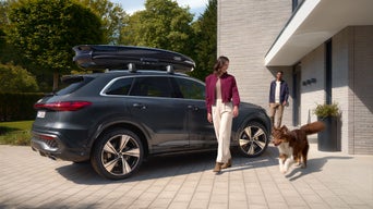 Ein grauer Audi Q5 mit montierter Dachbox steht vor einem modernen Haus, begleitet von einem Paar und ihrem Hund.