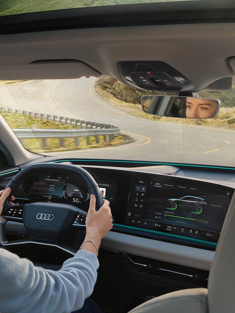 Eine junge Frau fährt einen Audi Q6 SUV e-tron, zu sehen sind das Cockpit, die kurvige Landstraße und ihr Blick im Rückspiegel.