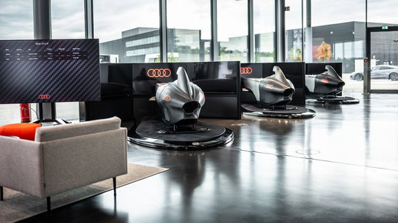 Drei Audi racing Simulator stehen im Showroom; der vordere ist unscharf, Audi Ringe leuchten auf den Trennwänden.