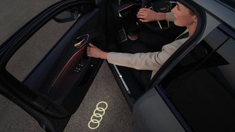 Eine elegant gekleidete Frau öffnet die Fahrertür aus dem Audi Q8 SUV e-hybrid heraus und dabei fällt das Audi Logo als Lichtsignatur auf die Straße.