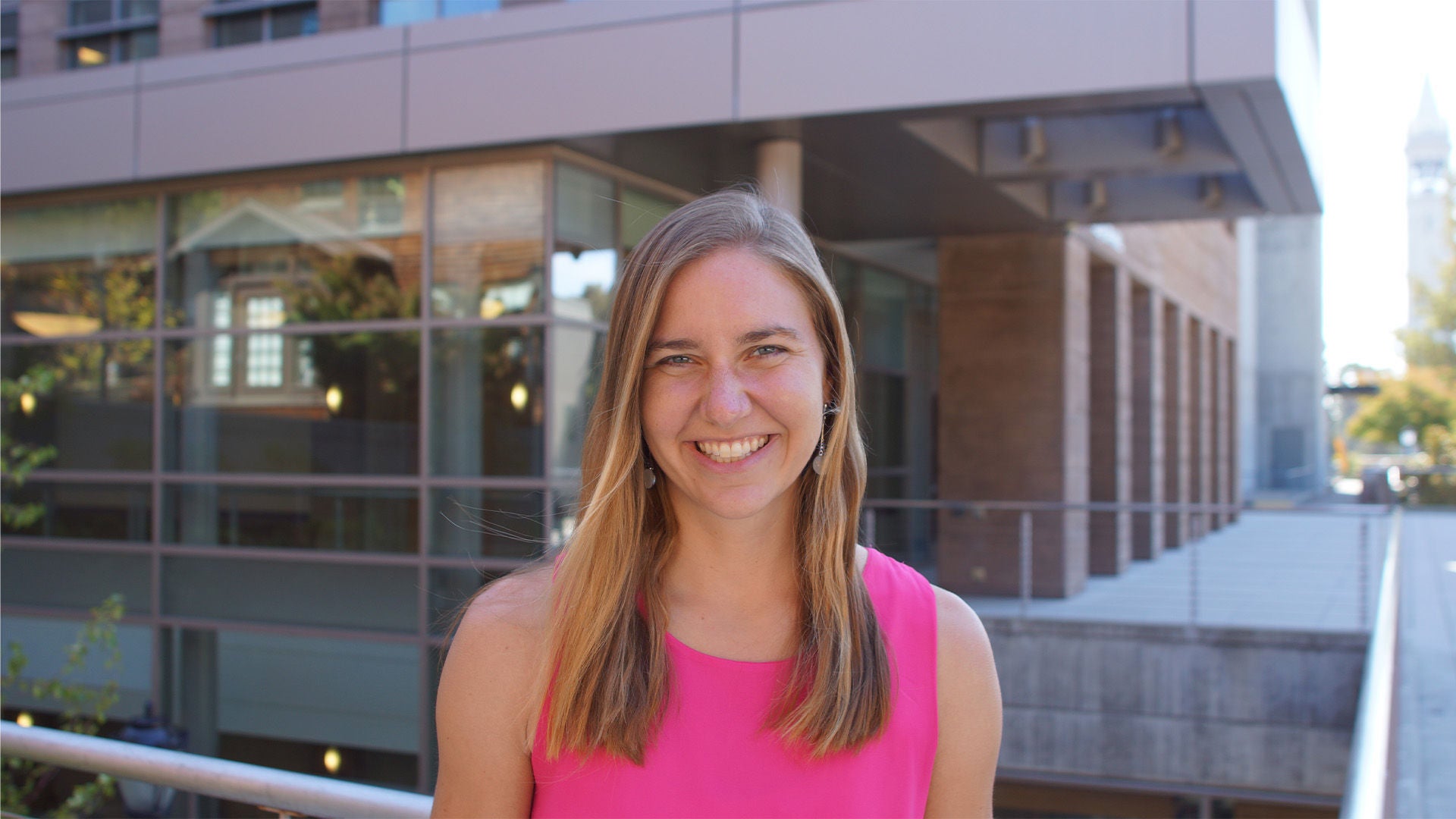 Frau mit langen blonden Haaren im pinken ärmellosen Oberteil, lächelnd, vor moderner Glasfassade.