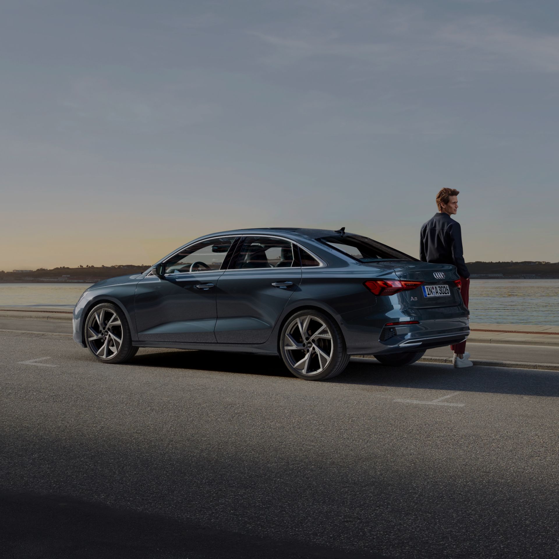 A person standing next to a blue sedan by a waterfront at dusk.