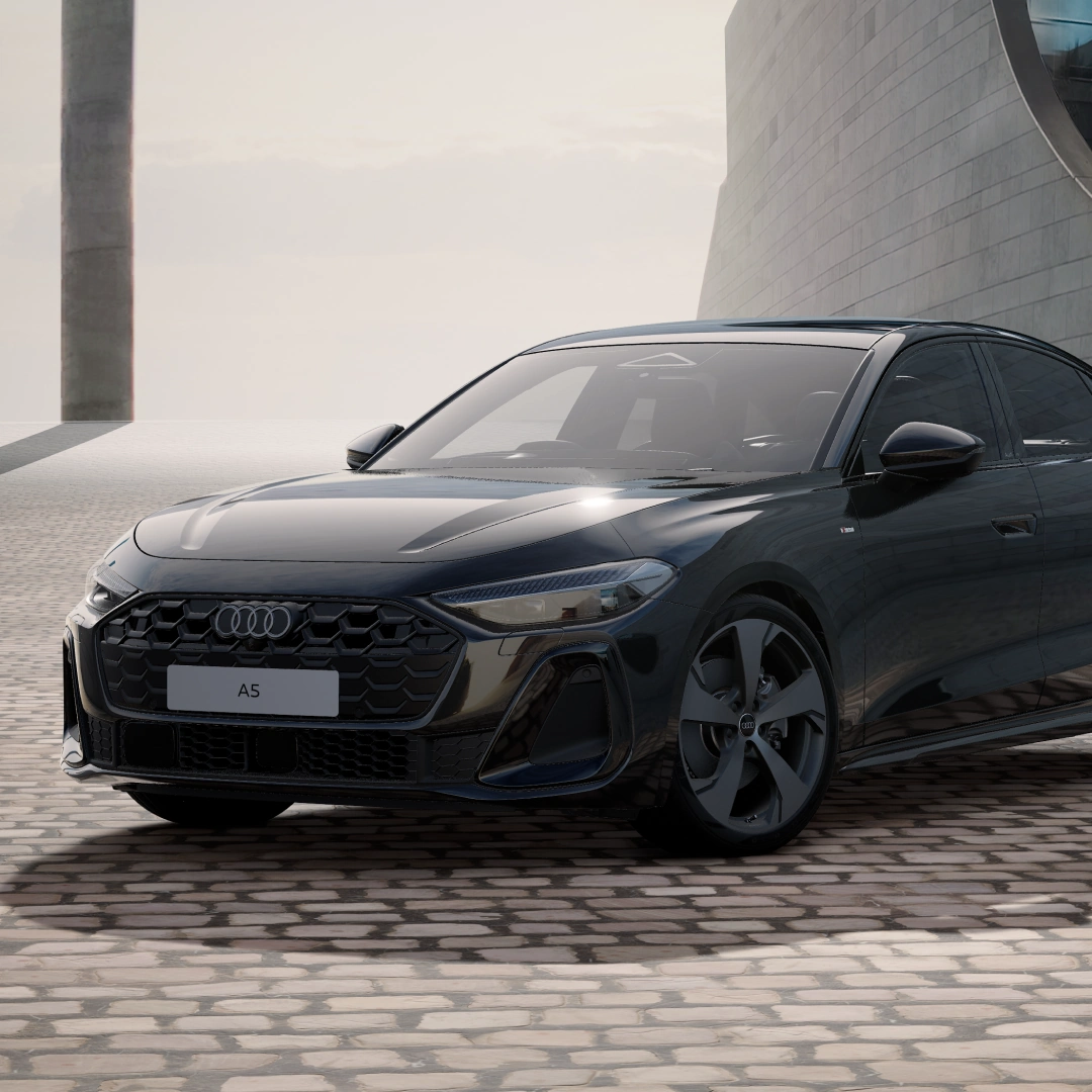Front three-quarter view of a dark blue Audi A5 parked on a cobbled surface beside a modern building, featuring a black singleframe grille, LED headlights and five-spoke aero-style alloy wheels.