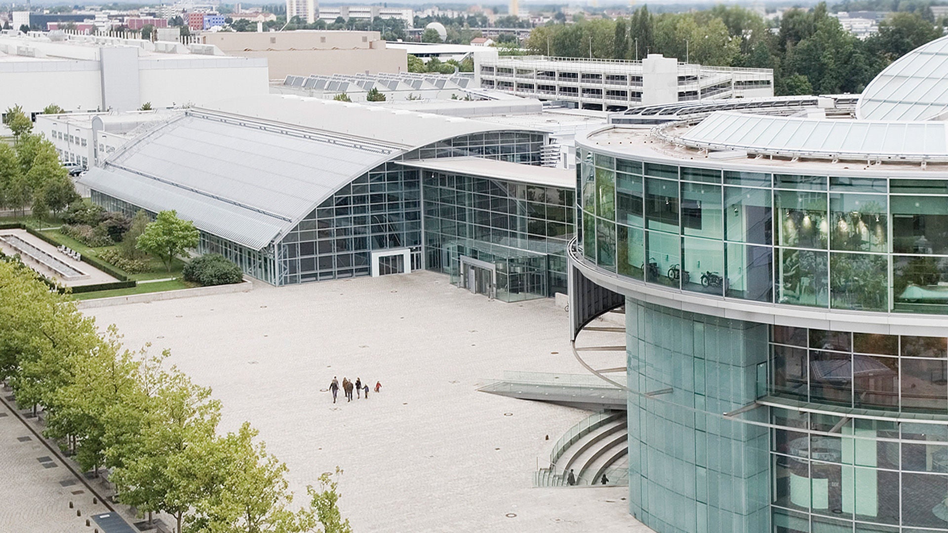 Aerial view of a modern glass and hall complex with a large forecourt; a few people are walking across the square.