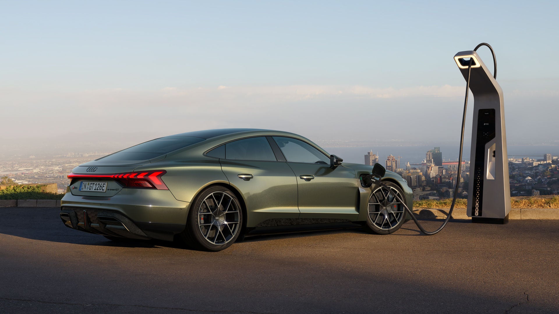 Grüner Audi RS e-tron GT performance lädt an einer Schnellladestation mit Stadtblick im Hintergrund.