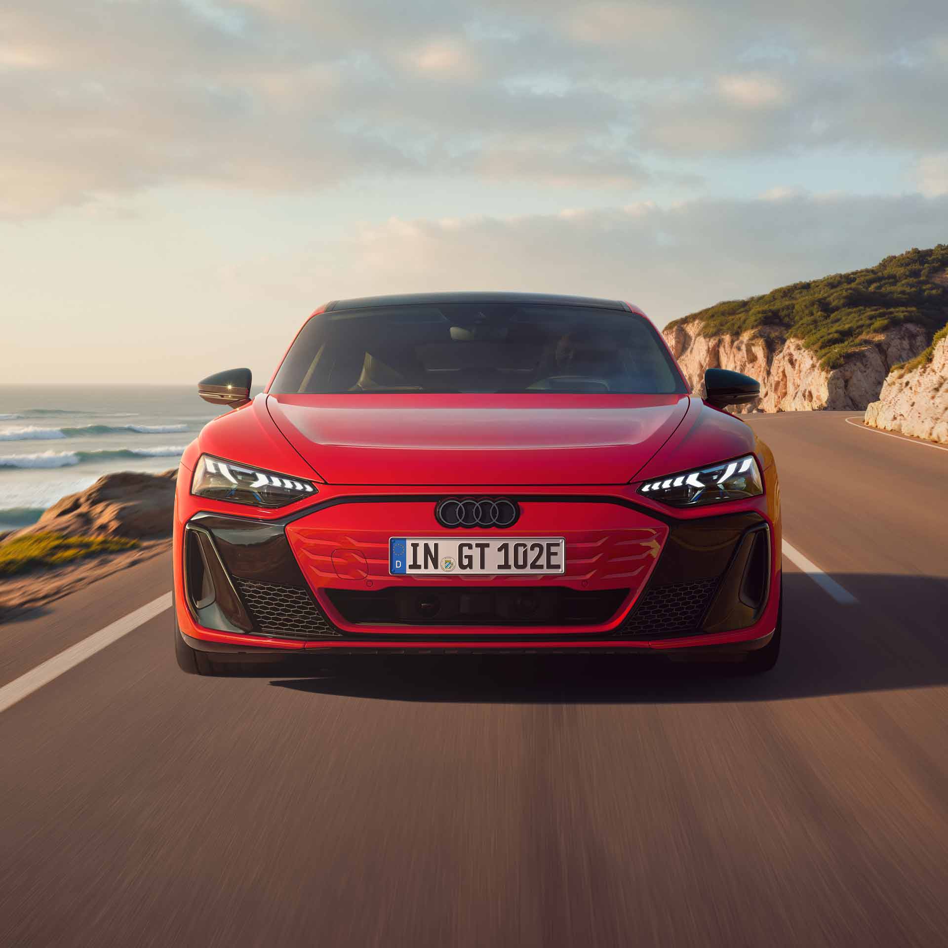 Frontansicht eines roten Audi e-tron GT quattro, der auf einer Küstenstraße fährt. Im Hintergrund sind Klippen, das Meer und der Himmel zu erkennen. Die gesamte Szenerie ist in das Licht eines Sonnenuntergangs gehüllt.