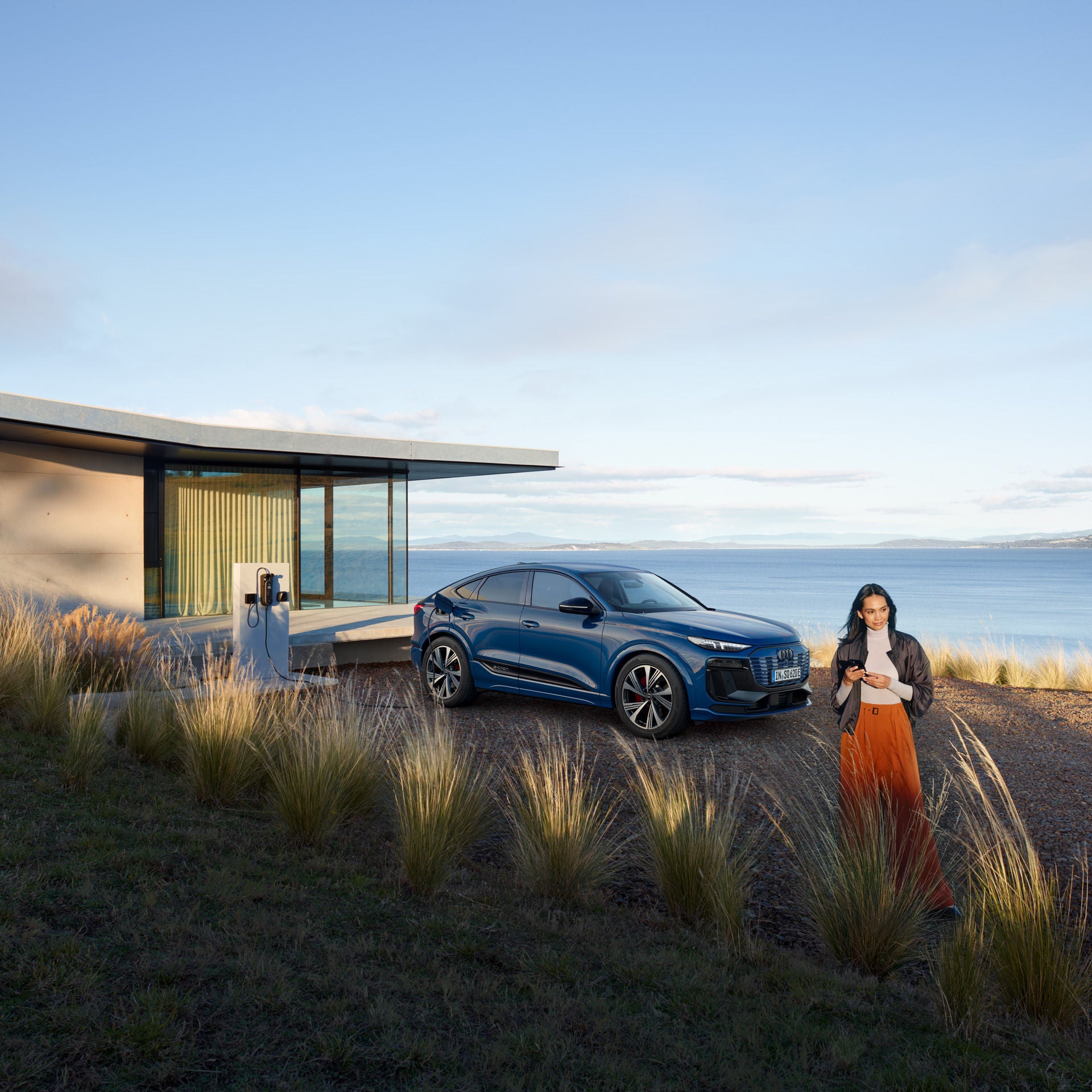 Ein blaues Audi Elektrofahrzeug steht an einer Ladestation neben einem modernen Haus mit großen Glasflächen am Meer. Im Vordergrund geht eine Frau mit Smartphone durch hohes Gras.