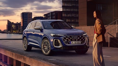 Der SQ5 SUV steht vor der Skyline einer Küstenstadt. Davor steht eine junge, modisch gekleidete Frau.
