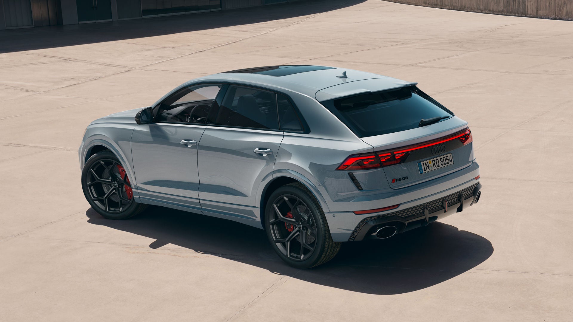 De RS Q8 SUV Performance met Audi exclusieve custom lak in Arrow Grey Pearl Effect, van achteren gezien vanuit een hoek op een zonovergoten parkeerplaats voor een modern gebouw.