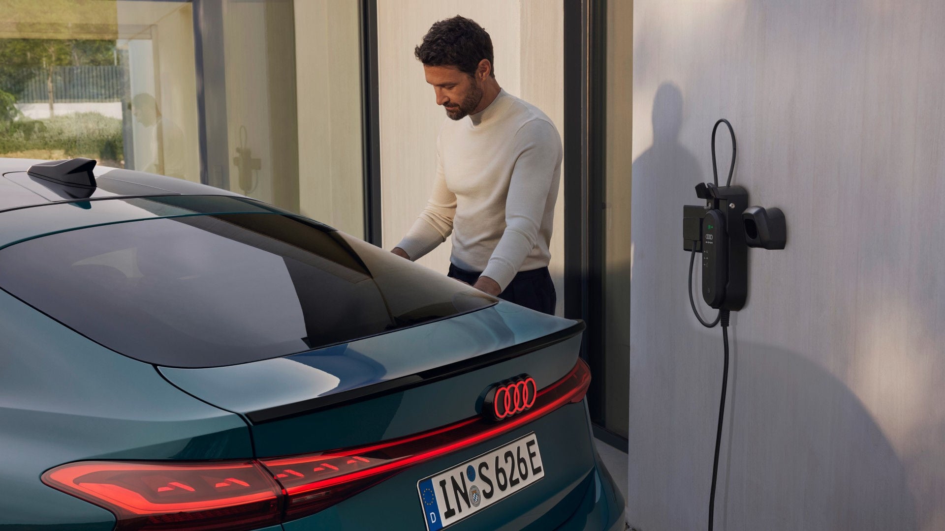 A man in a white sweater stands next to a teal Audi electric car parked in a modern garage. The car is plugged into a charger, suggesting charging use.