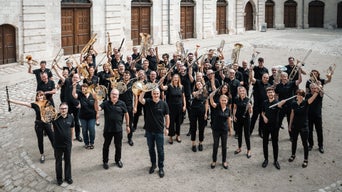 Wind band in black clothing stands outside and raises instruments in the air.
