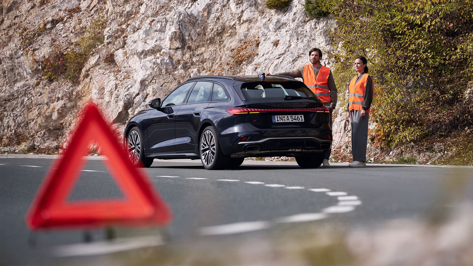 Un couple vêtu de gilets de sécurité orange se tient à côté de son Audi noire, au bord d'une route départementale. Ils sont en panne et, au premier plan, on aperçoit un triangle de signalisation rouge posé sur la chaussée.
