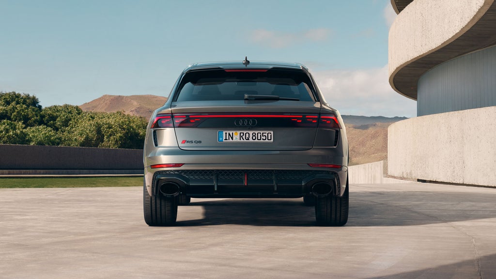 Der RS Q8 SUV von hinten parkend neben einem futuristischen Gebäude. Eine Mauer begrenzt das Grundstück, dahinter sind Wald und karge Gebirgszüge zu erkennen.
