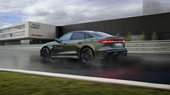 Grüne Audi RS 5 Limousine fährt auf nasser Strecke vor Audi-Gebäude, Rücklichter leuchten.