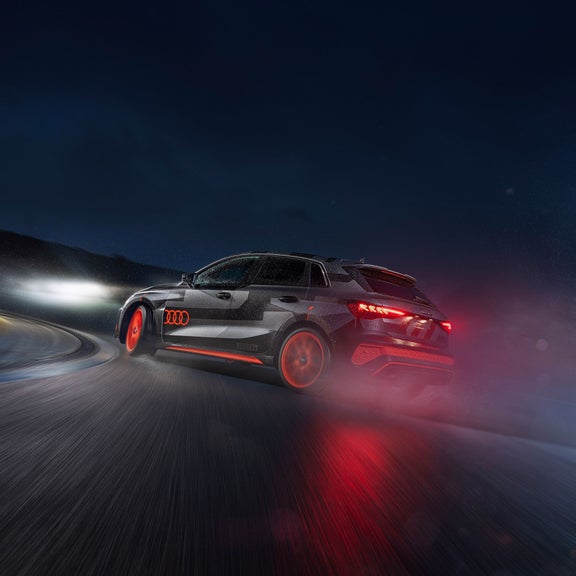 Schwarz/grauer Audi RS 3 driving experience mit rot leuchtenden Rücklichtern fährt bei Nacht durch eine nasse Kurve, Lichtspuren sichtbar.  