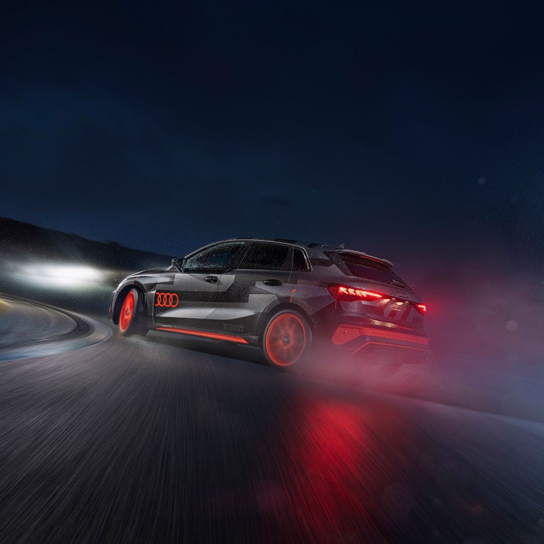 Schwarz/grauer Audi RS 3 driving experience mit rot leuchtenden Rücklichtern fährt bei Nacht durch eine nasse Kurve, Lichtspuren sichtbar.