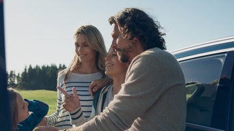 A happy family stands by a car in a sunny field, smiling and talking. The mood is joyful and relaxed, with a backdrop of trees and clear blue sky.