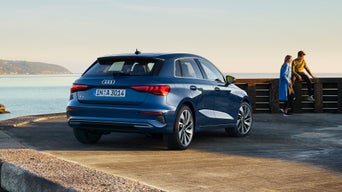 Blauer Audi A3 Sportback parkt am Wasser, zwei Personen stehen und sitzen an einer Holzbrüstung bei Sonnenuntergang