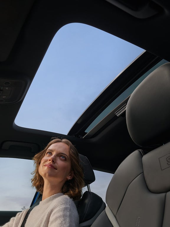 Frau mit welligem mittelbraunen Haar blickt durch geöffnetes Panoramadach im Audi Q2, schwarzer S-Sitz im Vordergrund.