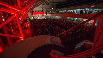 Konzertveranstaltung mit großem Publikum in modernem Saal mit roter Beleuchtung und Blick von einer Treppe.
