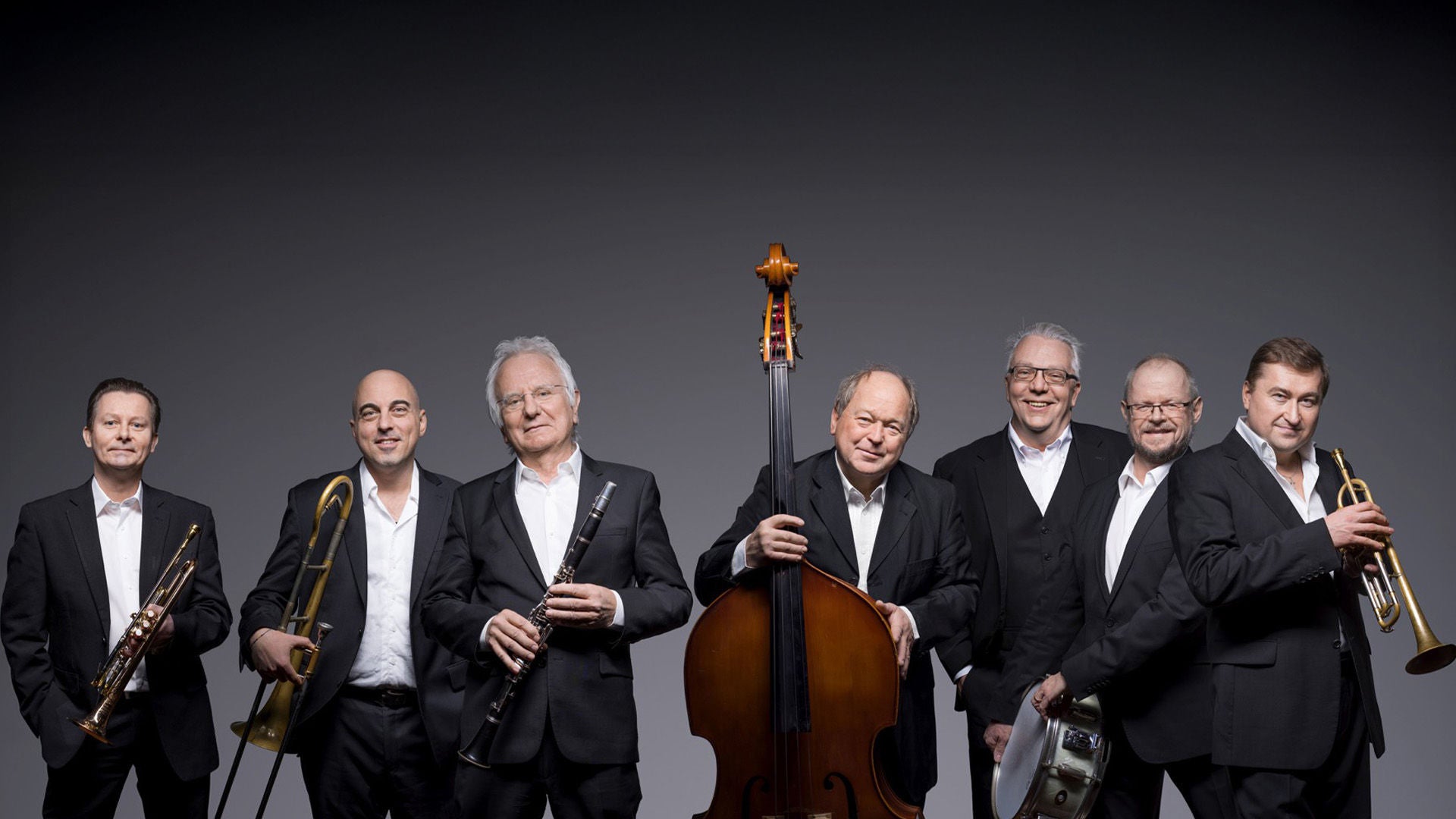 Sieben Musiker in schwarzen Anzügen stehen mit Trompete, Posaune, Klarinette, Kontrabass und Schlagzeug vor grauem Hintergrund.