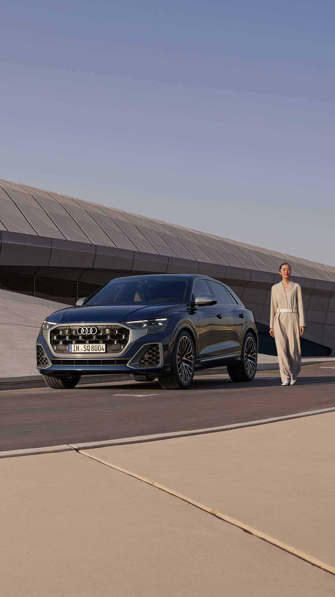 Eine Frau steht neben einem geparkten, dunkelblauen Audi SQ8 SUV vor einem modernen Gebäude.