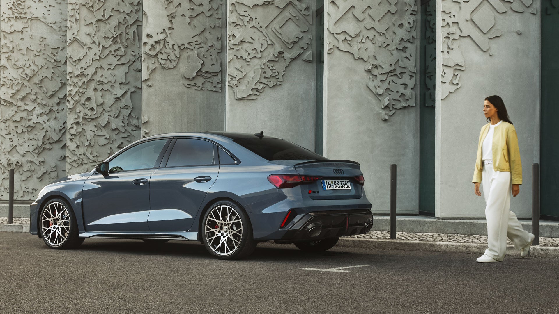 Frau geht an einer grauen Audi RS 3 Limousine vorbei, die vor einer strukturierten Betonwand geparkt ist.